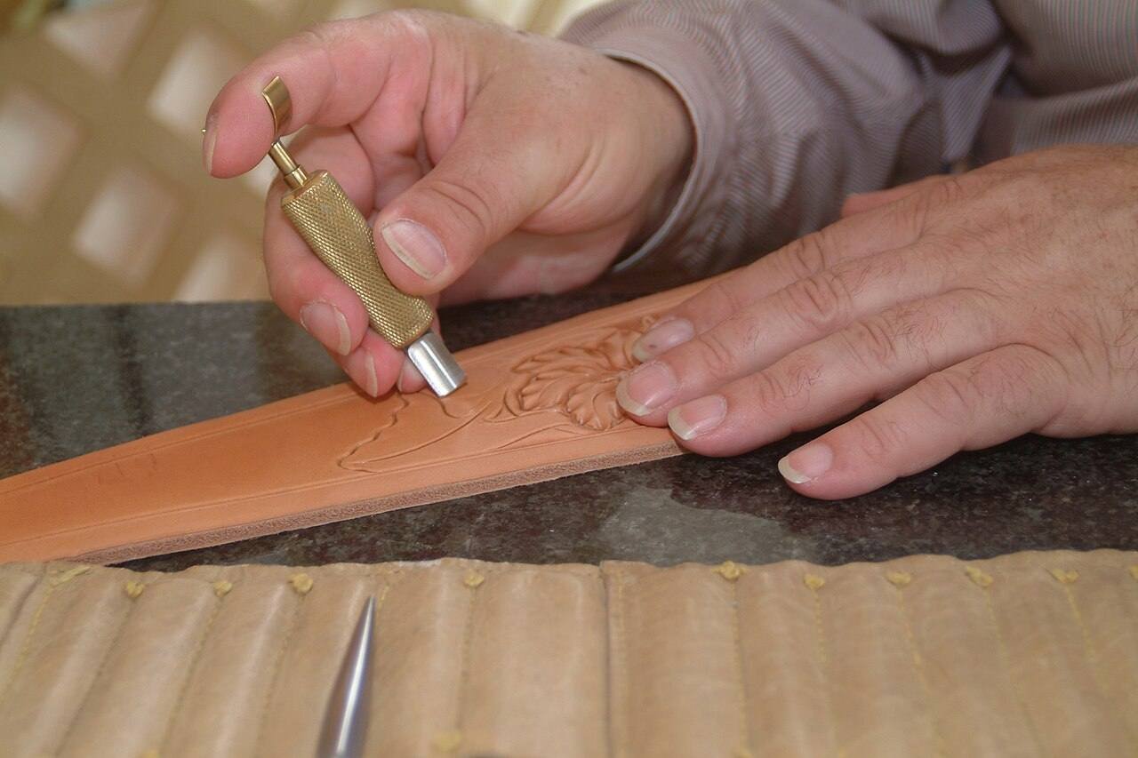 Mains d'un sellier ciselant un motif floral dans une bande de cuir naturel avec un couteau rotatif